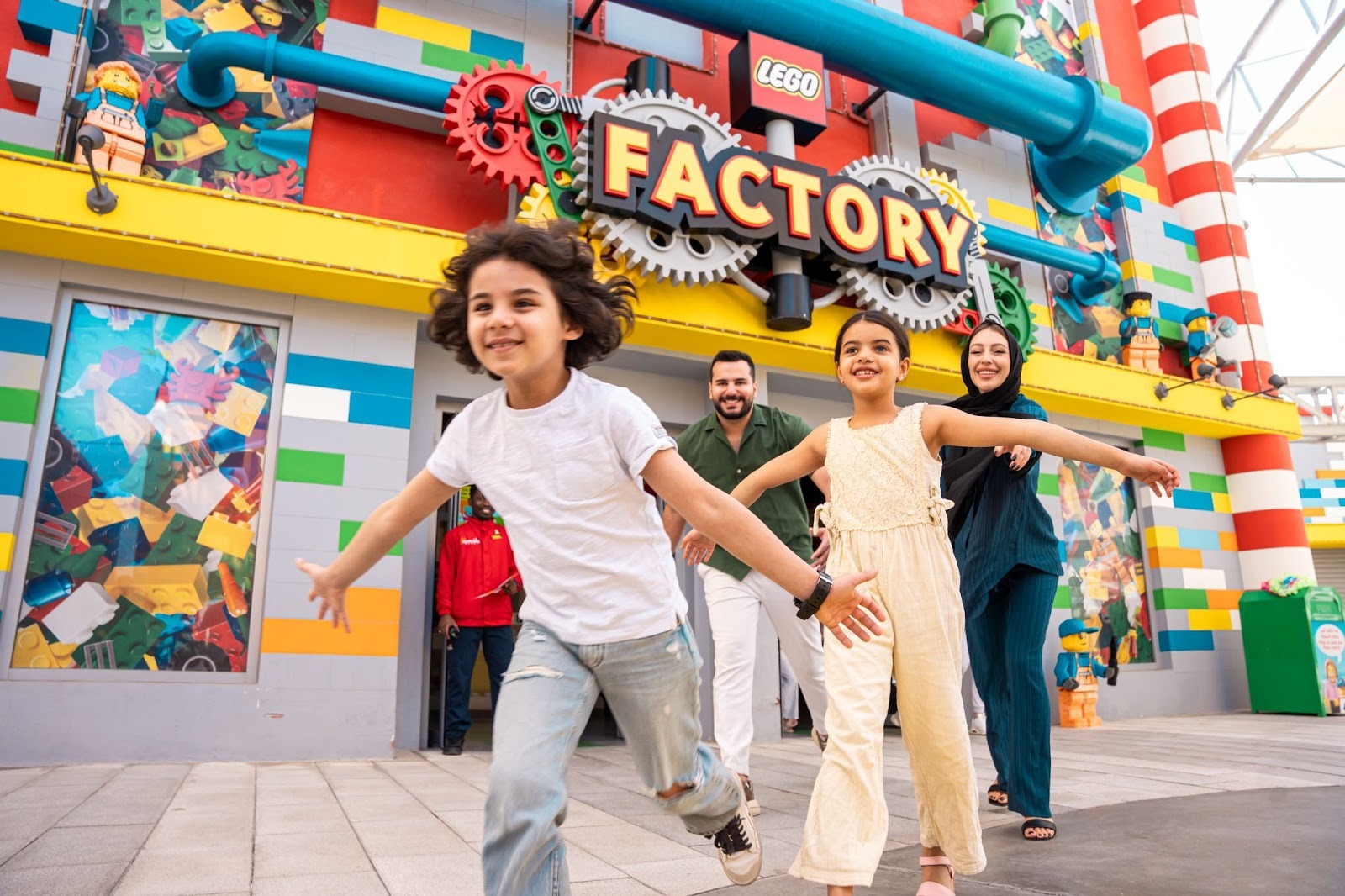A joyful family runs excitedly towards the camera in front of a colorful LEGO Factory entrance.