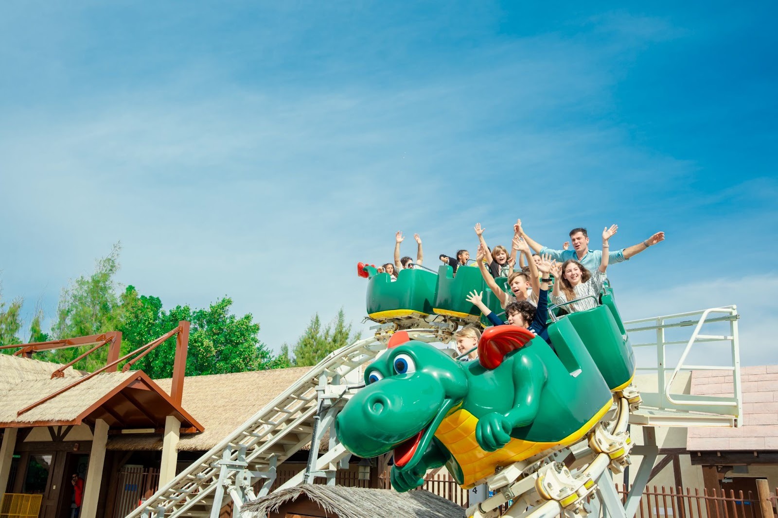 Group of people enjoy a ride on a dragon-themed roller coaster under a clear blue sky, hands raised in excitement